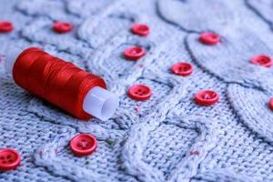 Beautiful texture of a soft warm natural sweater, fabrics with a knitted pattern of yarn and red small round buttons for sewing and a skein of red thread. The background photo