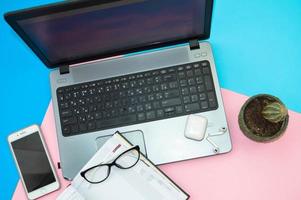 lugar de trabajo en casa, oficina en casa para trabajo remoto con una computadora portátil, teléfono móvil, teléfono inteligente, bloc de notas, auriculares y anteojos en un fondo rosa y azul foto