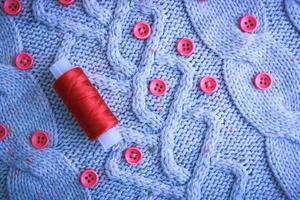 Beautiful texture of a soft warm natural sweater, fabrics with a knitted pattern of yarn and red small round buttons for sewing and a skein of red thread. The background photo