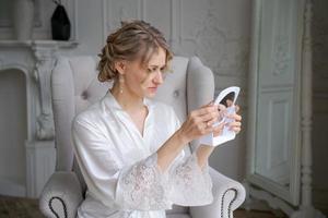 Beautiful blond woman, wearing white silk bathrobe, holding mirror and sitting