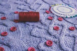 Beautiful texture of a soft warm natural sweater, knitted fabrics and red small round buttons for sewing and a skein of thread, needle bed. Flat lay. The background photo