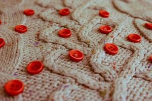 Beautiful texture of a soft warm natural sweater with a knitted pattern of yarn and red small round buttons for sewing. The background photo