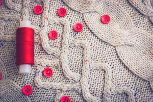 Beautiful texture of a soft warm natural sweater, fabrics with a knitted pattern of yarn and red small round buttons for sewing and a skein of red thread. The background photo