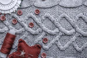 Beautiful texture of a soft warm natural sweater, fabrics with a knitted pattern and red small round buttons for sewing and a skein of thread, needle bed and needle pad. Flat lay. The background photo