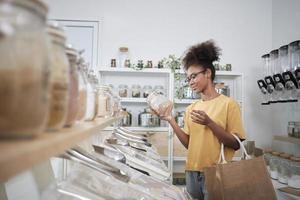 Young African American woman is choosing and shopping for organic products in refill store with reusable bag, zero-waste grocery, and plastic-free, eco environment-friendly, sustainable lifestyles. photo