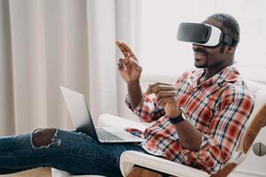 African american man in virtual reality glasses works or play video game on laptop, sitting in chair photo