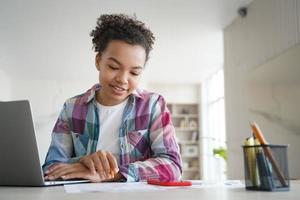 Una estudiante de raza mixta estudia sentada en un escritorio con una laptop haciendo los deberes. educación a distancia foto