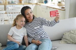 mamá feliz, la pequeña hija se toma una selfie o hace una videollamada, sostiene el teléfono, se sienta en el sofá foto
