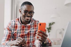 Modern african american man in glasses uses mobile apps, holding smartphone, sitting at desk smiling photo