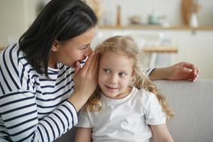 Mother telling secret, whispering to kid daughter, gossiping together at home. Trustful conversation photo