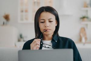 Puzzled businesswoman frowning works on laptop at home, solving problem, looking at computer screen photo