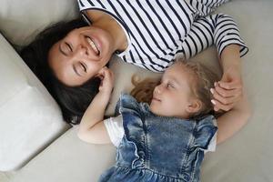 Little child daughter tickling laughing mother, lying on sofa, having fun together. Happy motherhood photo