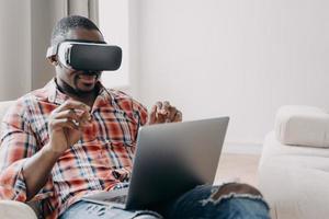 African american man software developer wearing virtual reality glasses works, creates app at laptop photo