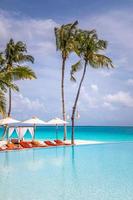 hermoso panorama de piscina de playa vertical. palmeras, sillas de playa, camas con piscina infinita cerca del mar y el horizonte. fondo de verano tranquilo, paisaje recreativo de ocio de viaje. balneario tropical foto