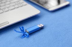 Brilliant blue usb flash memory card with a blue bow lies on a blanket of soft and furry light blue fleece fabric beside to a white laptop and smartphone. Classic female gift design for a memory card photo