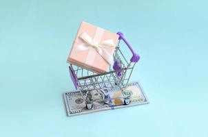 Gift box in a small shopping cart lies on a dollar bills on light blue background photo