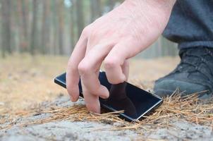 Male hand picking up lost mobile phone from a ground in autumn fir wood path. The concept of finding a valuable thing and good luck photo