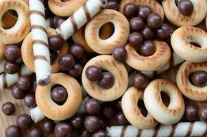 A lot of crispy sweet tubules, chocolate melting balls and yellow bagels lie on a wooden surface. Close up view photo