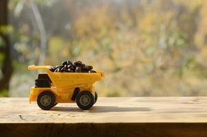 A small yellow toy truck is loaded with brown coffee beans. A car on a wooden surface against a background of autumn forest. Extraction and transportation of coffee photo