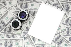 Two photographic lenses and white notebook lie on the background of a lot of dollar bills. Space for text photo