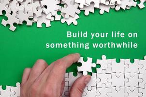 Build your life on something worthwhile. The hand folds a white jigsaw puzzle and a pile of uncombed puzzle pieces lies against the background of the green surface photo