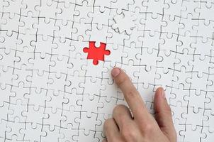 The texture of a white jigsaw puzzle in the assembled state with one missing element, forming a red space, pointed to by the finger of the male hand photo