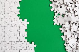 Fragment of a folded white jigsaw puzzle and a pile of uncombed puzzle elements against the background of a green surface. Texture photo with space for text