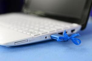 A brilliant blue USB flash drive with a blue bow is connected to a white laptop, which lies on a blanket of soft and fluffy light blue fleece fabric. Classic female design for a memory card photo