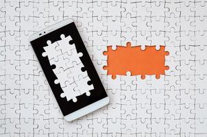 A modern big smartphone with several puzzle elements on the touch screen lies on a white jigsaw puzzle in an assembled state with missing elements photo