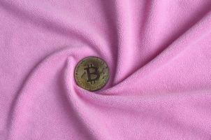 The golden bitcoin lies on a blanket made of soft and fluffy light pink fleece fabric with a large number of relief folds. The shape of the folds resembles a fan from a video card cooler photo