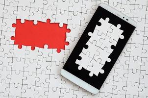 A modern big smartphone with several puzzle elements on the touch screen lies on a white jigsaw puzzle in an assembled state with missing elements photo
