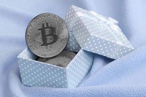 The silver bitcoin lies in a small blue gift box with a small bow on a blanket made of soft and fluffy light blue fleece fabric with a large number of relief folds photo