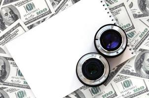 Two photographic lenses and white notebook lie on the background of a lot of dollar bills. Space for text photo