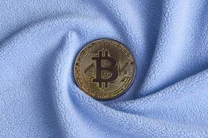 The golden bitcoin lies on a blanket made of soft and fluffy light blue fleece fabric with a large number of relief folds. The shape of the folds resembles a fan from a video card cooler photo