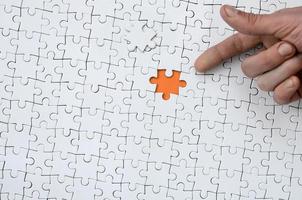 The texture of a white jigsaw puzzle in the assembled state with one missing element, forming an orange space, pointed to by the finger of the male hand photo