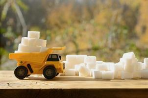 un pequeño camión de juguete amarillo está cargado con cubos de azúcar blanco cerca de los restos de azúcar. un coche sobre una superficie de madera sobre un fondo de bosque otoñal. extracción y transporte de azúcar foto
