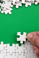 The hand folds a white jigsaw puzzle and a pile of uncombed puzzle pieces lies against the background of the green surface. Texture photo with space for text
