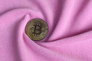 The golden bitcoin lies on a blanket made of soft and fluffy light pink fleece fabric with a large number of relief folds. The shape of the folds resembles a fan from a video card cooler photo
