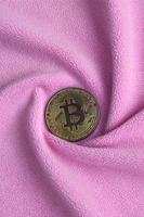 The golden bitcoin lies on a blanket made of soft and fluffy light pink fleece fabric with a large number of relief folds. The shape of the folds resembles a fan from a video card cooler photo