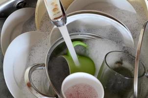 Dirty dishes and unwashed kitchen appliances lie in foam water under a tap from a kitchen faucet photo