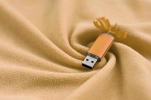 Orange usb flash memory card with a bow lies on a blanket of soft and furry light orange fleece fabric with a lot of relief folds. Memory storage device in women's design photo