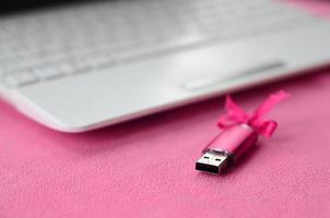 Brilliant pink usb flash memory card with a pink bow lies on a blanket of soft and furry light pink fleece fabric beside to a white laptop. Classic female gift design for a memory card photo