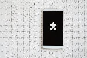 A modern big smartphone with several puzzle elements on the touch screen lies on a white jigsaw puzzle in an assembled state photo