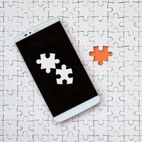 A modern big smartphone with several puzzle elements on the touch screen lies on a white jigsaw puzzle in an assembled state with missing elements photo