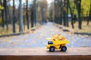 A small toy yellow truck is loaded with yellow fallen leaves. The car stands on a wooden surface against a background of a blurry autumn park. Cleaning and removal of fallen leaves. Seasonal works photo