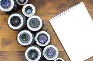 Several photographic lenses and white notebook lie on a brown wooden background. Space for text photo