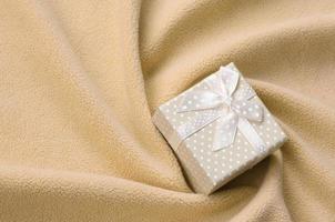 A small gift box in orange with a small bow lies on a blanket of soft and furry light orange fleece fabric with a lot of relief folds. Packing for a gift to your lovely girlfriend photo