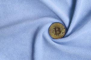 The golden bitcoin lies on a blanket made of soft and fluffy light blue fleece fabric with a large number of relief folds. The shape of the folds resembles a fan from a video card cooler photo