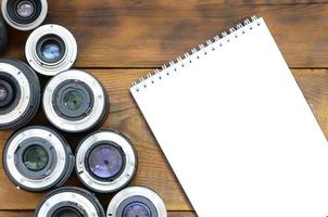 Several photographic lenses and white notebook lie on a brown wooden background. Space for text photo