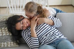 pequeña hija feliz haciéndole cosquillas a mamá o niñera, sentada en el sofá, divirtiéndose juntos foto